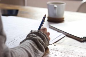 Person's hand writing with a blue pen on a white paper, coffee mug in background.