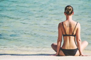 Woman in black bikini meditating on a beach, back view showing a back tattoo.