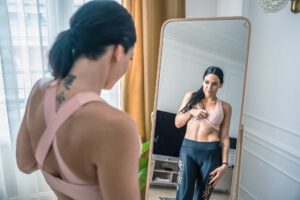 Woman admiring her physique in a mirror, wearing a sports bra and leggings.