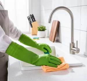 Person wearing green cleaning gloves washes dishes in a white sink, using a sponge and cloth.